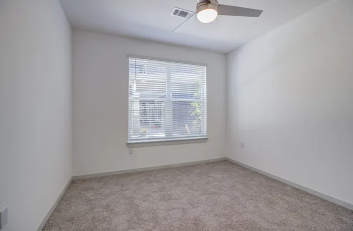 Unfurnished apartment bedroom with carpet, large window, ceiling fan, and overhead lighting at Garrett Forum in Fort Myers, Florida.
