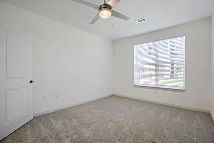 Bright carpeted bedroom with ceiling fan and large window with blinds at Garrett Harvest Junction West apartments in Longmont, CO