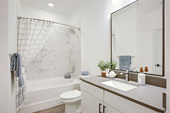 Staged full bathroom with marble-look tile tub surround, white vanity, framed mirror, and gray countertop at Garrett Harvest Junction West in Longmont, CO
