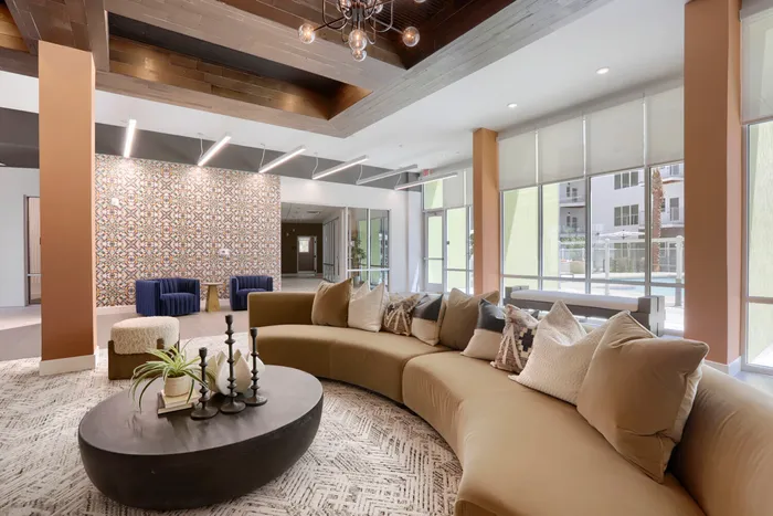Garrett Uptown clubhouse lounge with curved camel sectional sofa, round coffee table, decorative tile accent wall, and floor-to-ceiling windows in Chandler, AZ