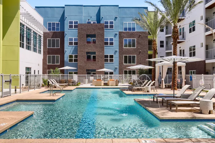 Garrett Uptown pool deck with lounge chairs, umbrellas, fire pit seating area, water feature jets, and colorful apartment building exterior in Chandler, AZ