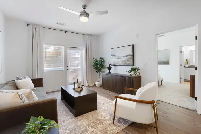 Garrett Uptown B2 apartment living room with leather sofa, wood coffee table, ceiling fan, patio doors, and en suite bedroom view in Chandler, AZ