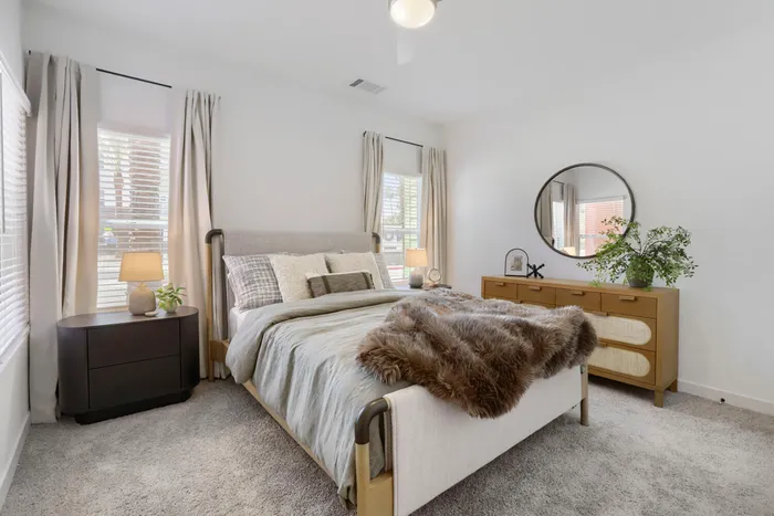 Garrett Uptown B2 apartment second bedroom with plush carpet, upholstered bed, walnut dresser, round mirror, and dual windows in Chandler, AZ