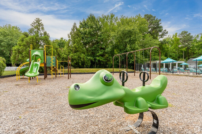 Inviting playground with vibrant green alligator spring rider, slide, and swings, nestled by lush trees and adjacent to a pool with umbrella-shaded seating.