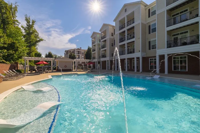 Luxurious apartment complex featuring a sparkling outdoor pool with refreshing fountains, cozy loungers, and elegant architecture under a sunny sky.