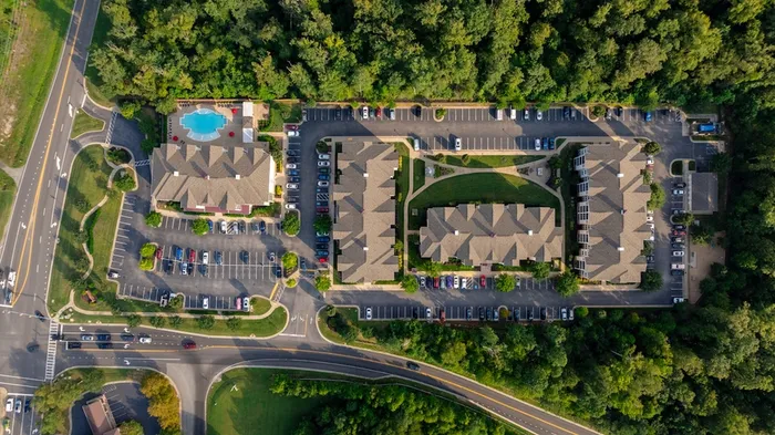 Aerial view of a vibrant apartment complex surrounded by lush greenery, featuring ample parking and a large, inviting swimming pool.