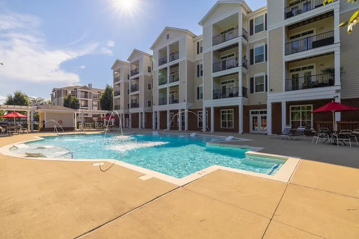 Luxurious apartment complex with sunlit swimming pool, elegant balconies, and red parasols creating a resort-style ambiance.