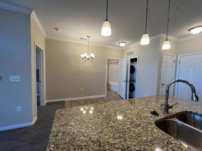 Modern apartment interior featuring a granite kitchen countertop, elegant pendant lighting, and an open layout leading to a cozy room with a washer and dryer.