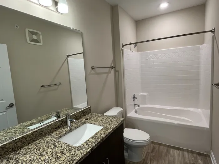 Modern bathroom featuring elegant granite countertops, a spacious mirror, and a pristine white tile bathtub.