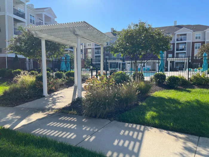 Charming apartment community showcasing a sunlit path under a white pergola, lush gardens, and a sparkling swimming pool.