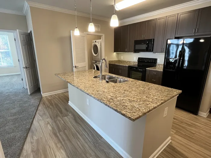 Stylish open-concept kitchen with a granite island, sleek appliances, and ample storage. Bright room with modern wood flooring and cozy carpeted bedroom visible.