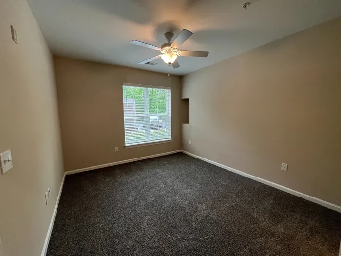 Bright bedroom with large window, plush carpet, and ceiling fan, offering a cozy and inviting atmosphere.