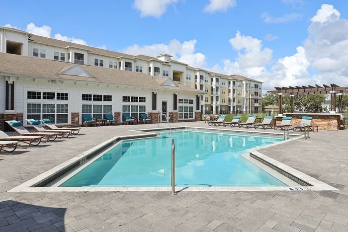 Luxurious apartment complex with a sparkling pool surrounded by sun loungers, elegant pergola, and stylish clubhouse under a bright blue sky.