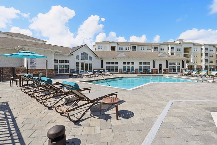 Luxurious apartment complex featuring a sparkling outdoor pool, surrounded by elegant lounging chairs and vibrant umbrellas against a bright blue sky.