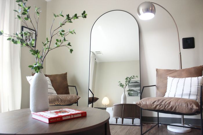 Modern apartment corner with elegant seating, an arched mirror, and stylish decor including a sleek floor lamp and a vase with green branches on a wooden table.