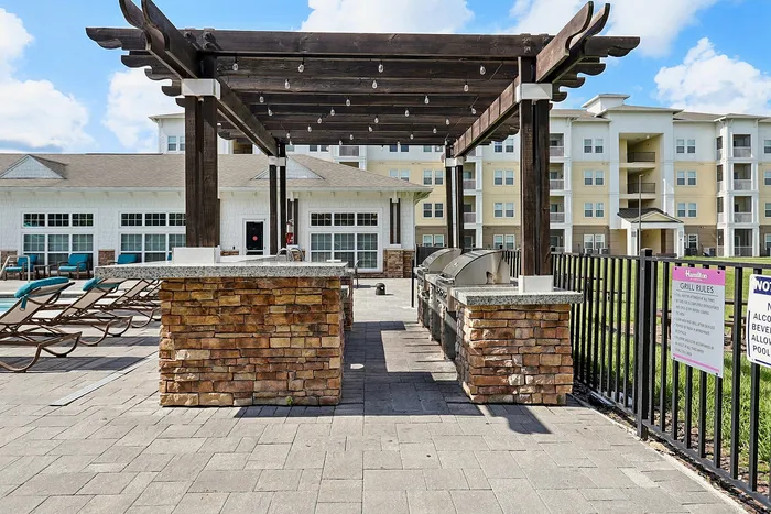 Charming outdoor area featuring a modern barbecue setup with stone accents under a stylish pergola, surrounded by cozy lounge chairs and a vibrant apartment complex.