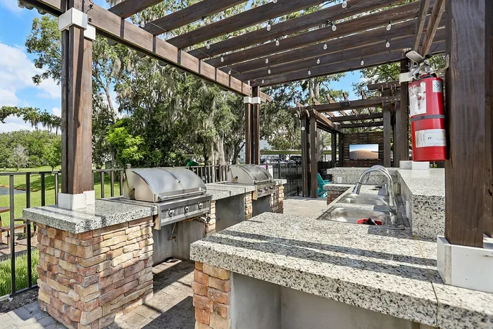 Charming outdoor kitchen with stone countertops and grills beneath a wooden pergola, surrounded by lush greenery.