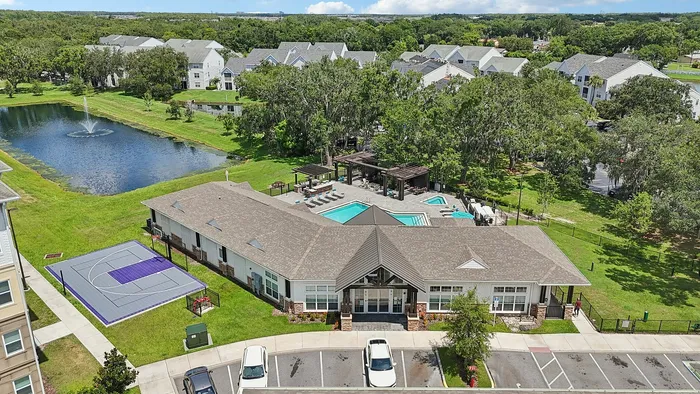 Aerial view of a vibrant apartment community featuring a sparkling swimming pool, modern sports court, serene pond with a fountain, and lush greenery, set against a backdrop of charming residential buildings.