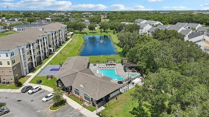Aerial view of a vibrant apartment complex featuring modern buildings, a scenic pond with a fountain, a sparkling swimming pool, and a basketball court, all surrounded by lush greenery.