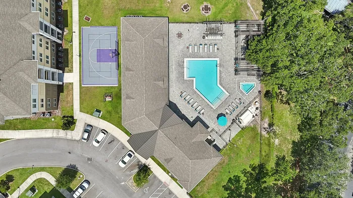 Aerial view of a luxurious apartment complex featuring a vibrant pool, sun loungers, and a modern basketball court surrounded by lush greenery.