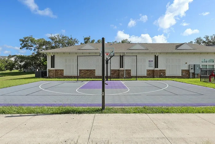 Modern outdoor basketball court with vibrant purple accents next to a charming brick and wood-paneled building under a clear blue sky.