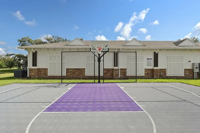 Modern outdoor basketball court with vibrant purple and gray flooring, nestled beside a charming suburban building under a bright blue sky.