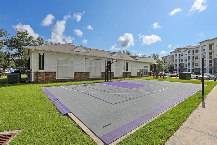 Modern residential complex featuring a vibrant outdoor basketball court with purple accents, elegant white-finished building, and lush green surroundings on a sunny day.
