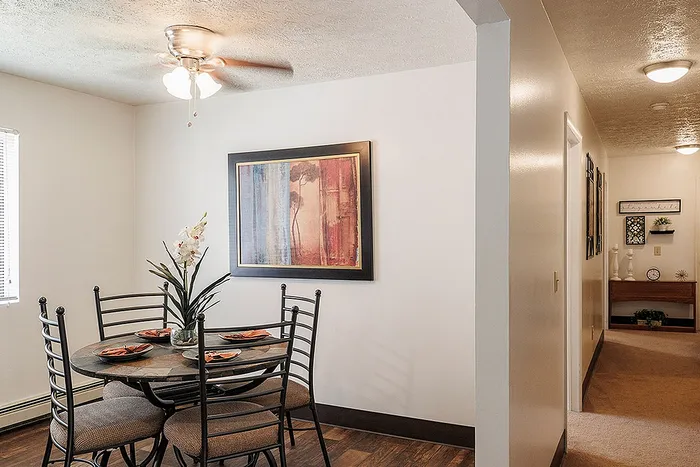 Charming dining area with modern decor, featuring a round table set for four under a stylish ceiling fan, adjacent to a bright hallway adorned with elegant artwork.
