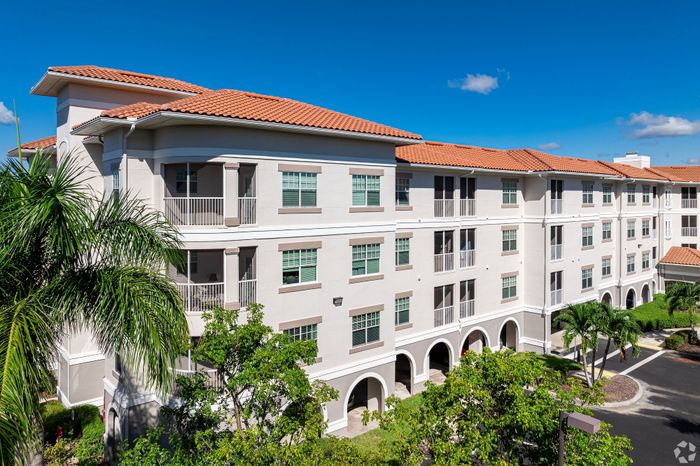 Modern Mediterranean-style apartment building with elegant arches, expansive balconies, and lush palm trees under a clear blue sky.