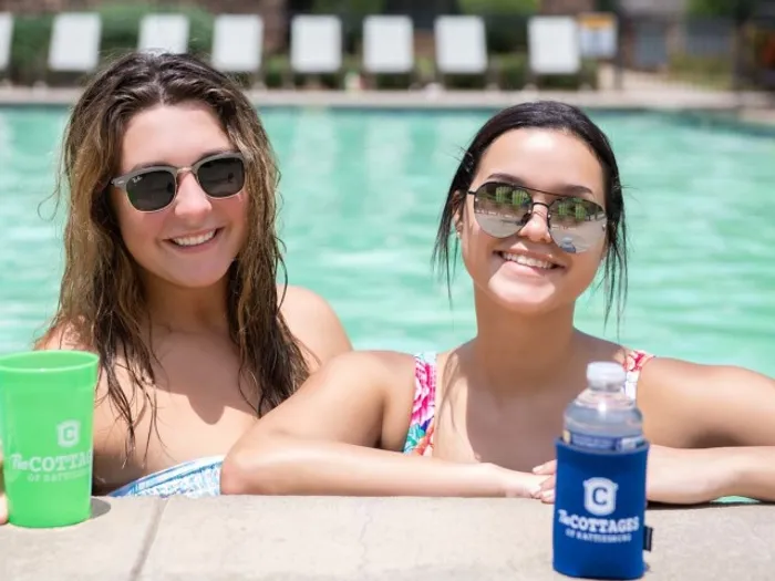 Two smiling residents relaxing in the pool at The Cottages of Hattiesburg, enjoying the sun and socializing with branded drinkware.