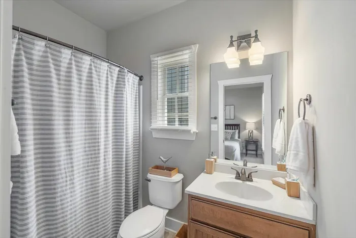 Clean and bright private bathroom at The Cottages of Hattiesburg, featuring a vanity, mirror, and shower with a striped curtain, offering a comfortable living space for residents.