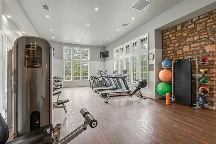 Fully equipped fitness center at The Cottages of Hattiesburg with cardio machines, strength equipment, and large windows providing natural light for a bright and spacious workout environment.