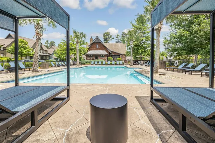 Poolside view of the resort-style pool at The Cottages of Hattiesburg with shaded lounge seating and palm trees.