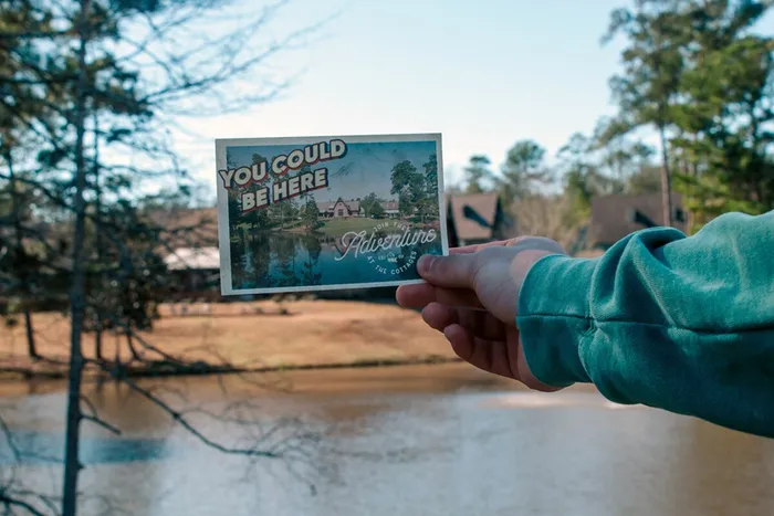 A hand holding a postcard with the message "You Could Be Here" in front of a scenic lake view at The Cottages of Hattiesburg, highlighting student living near Southern Miss.