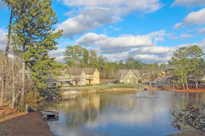 Beautiful view of The Cottages of Hattiesburg with a peaceful pond and fountain, surrounded by greenery and cottage-style townhomes.
