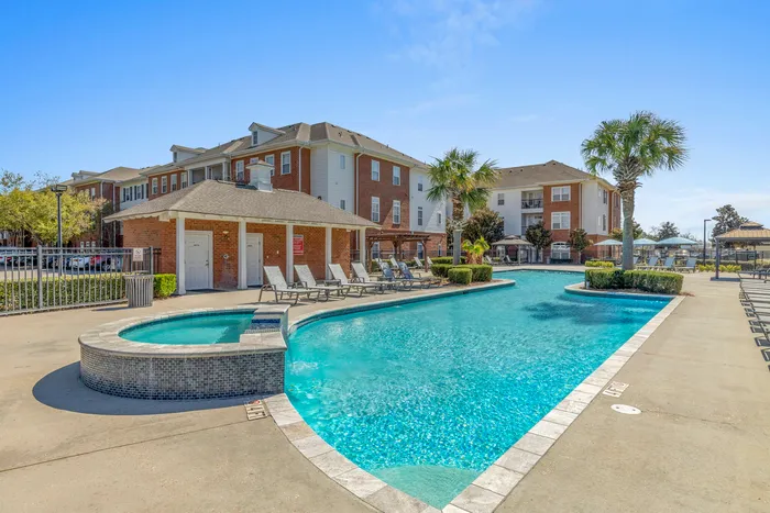Inviting apartment complex with a luxurious pool surrounded by sun loungers, vibrant landscaping, and stylish brick buildings under a clear blue sky.