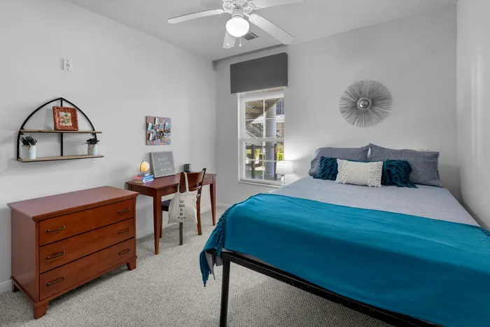 Bright and cozy bedroom featuring a stylish bed with blue accents, elegant wall art, and a functional workspace by a sunny window.