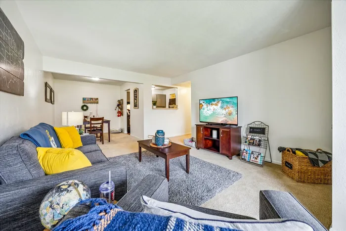 Inviting living room featuring a cozy gray sofa with bright yellow pillows, a modern coffee table, and a wall-mounted TV, leading into a dining area with elegant decor.
