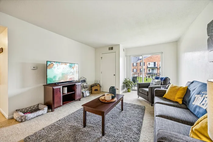 Bright and inviting living room featuring cozy gray sofas, large flat-screen TV, elegant wooden coffee table, and ample natural light from sliding glass doors with a view.