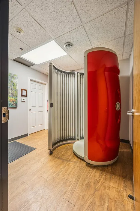 Modern space with sleek wooden flooring and a distinct red cylindrical stand up tanning bed, complemented by bright overhead lighting and contemporary decor.