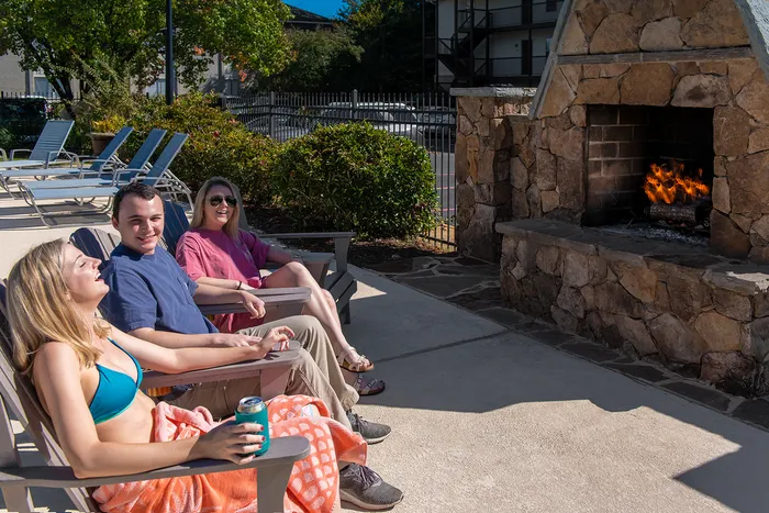 Relaxing outdoor lounge area with smiling friends by a warm stone fireplace, perfect for sunny social gatherings.