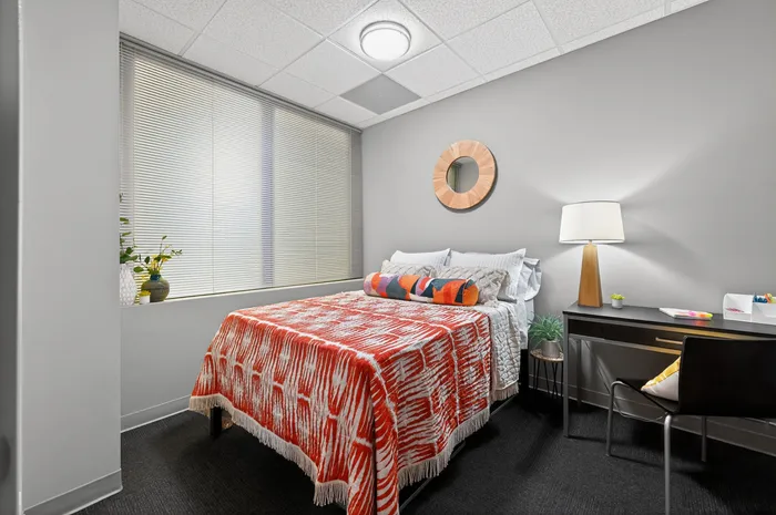 Modern bedroom with cozy double bed, vibrant orange-patterned cover, and sleek workspace under ambient lighting.
