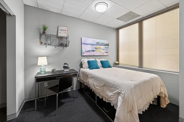 Cozy bedroom with modern decor, featuring a comfortable bed, stylish desk, and large window with blinds for natural light.