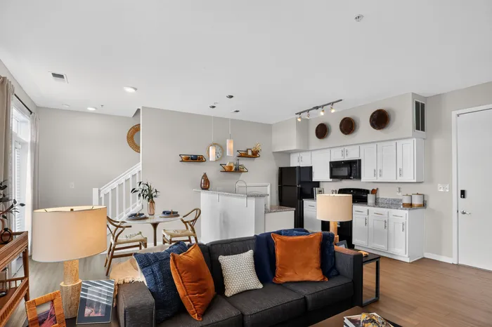 Modern open-plan living room and kitchen with stylish decor, showcasing sleek white cabinets, cozy seating with vibrant pillows, and elegant dining area.
