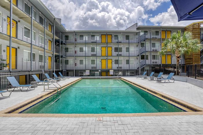 Modern apartment complex with a pristine swimming pool, surrounded by stylish lounge chairs and vibrant yellow accents, under a bright, cloudy sky.