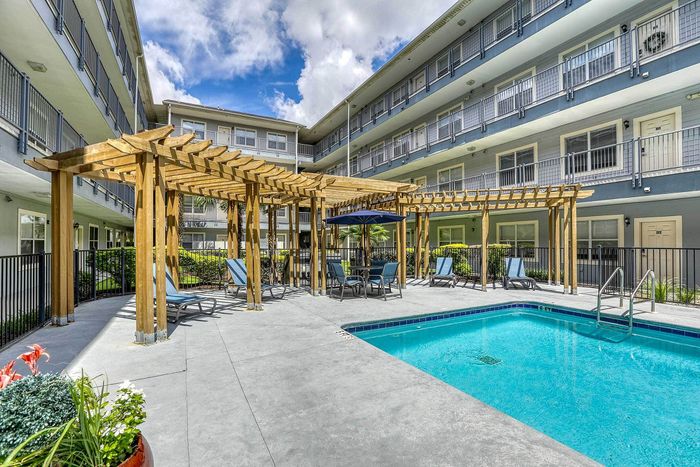 Charming apartment complex courtyard featuring a pristine swimming pool, wooden pergola, and relaxing lounge chairs under a bright blue sky.