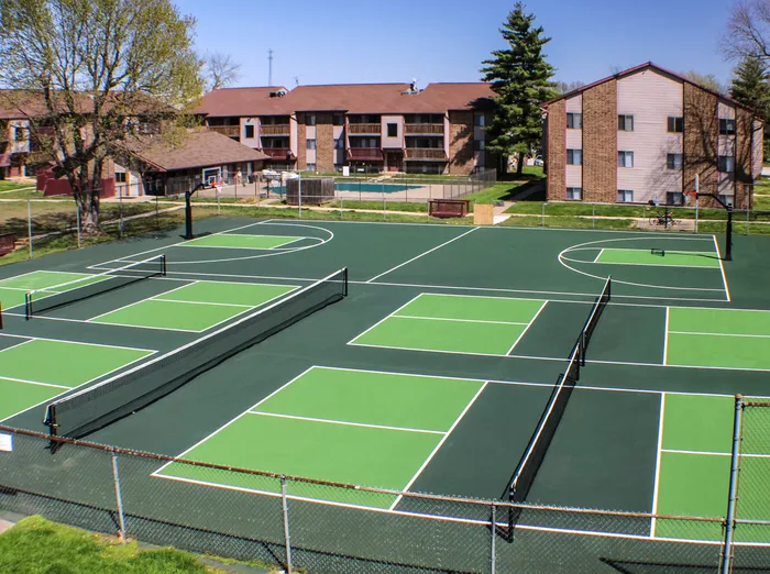Outdoor recreational area featuring multiple pickleball courts and a basketball court, surrounded by a chain-link fence and landscaped greenery, with apartment buildings in the background.