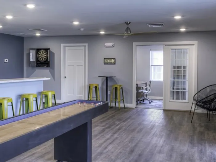 Community game area with shuffleboard &ndash; Spacious clubhouse game room with shuffleboard table, dart board, and bright overhead lighting at The Field Apartments.