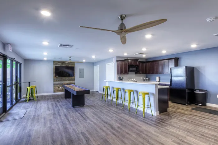Clubhouse with open seating and kitchen &ndash; Spacious clubhouse lounge featuring wood-style flooring, a large flat-screen TV, shuffleboard table, and modern kitchen with green bar stools and stainless steel appliances.