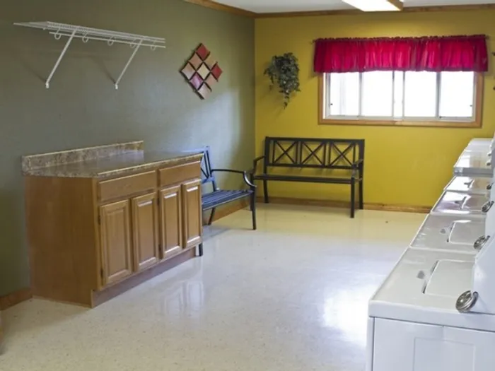 Laundry room with green and yellow walls, featuring a wooden counter, shelving, a bench under a window with a red curtain, and a washing machine.